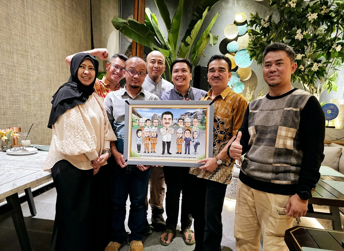 Tujuh rekan kerja berkumpul di ruang tunggu kantor sambil memegang karikatur kelompok berbingkai bertuliskan ucapan terima kasih untuk atasan mereka