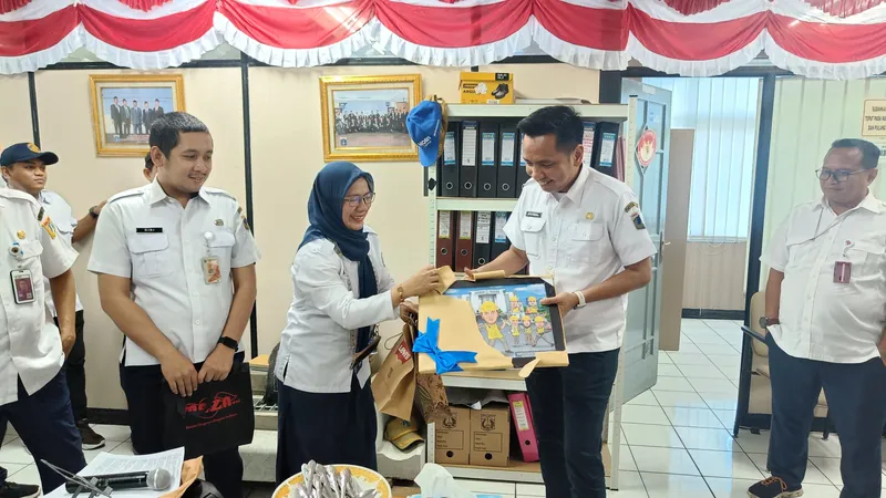 Serah terima karikatur berbingkai antara dua pejabat berseragam putih disaksikan rekan sekantor.