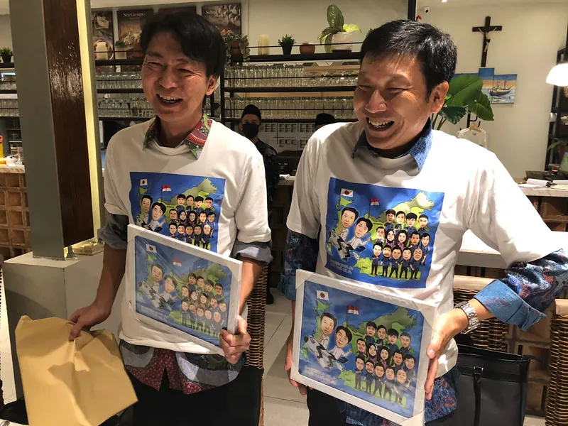 Dua pria tertawa sambil masing-masing memegang karikatur kelompok berbingkai.