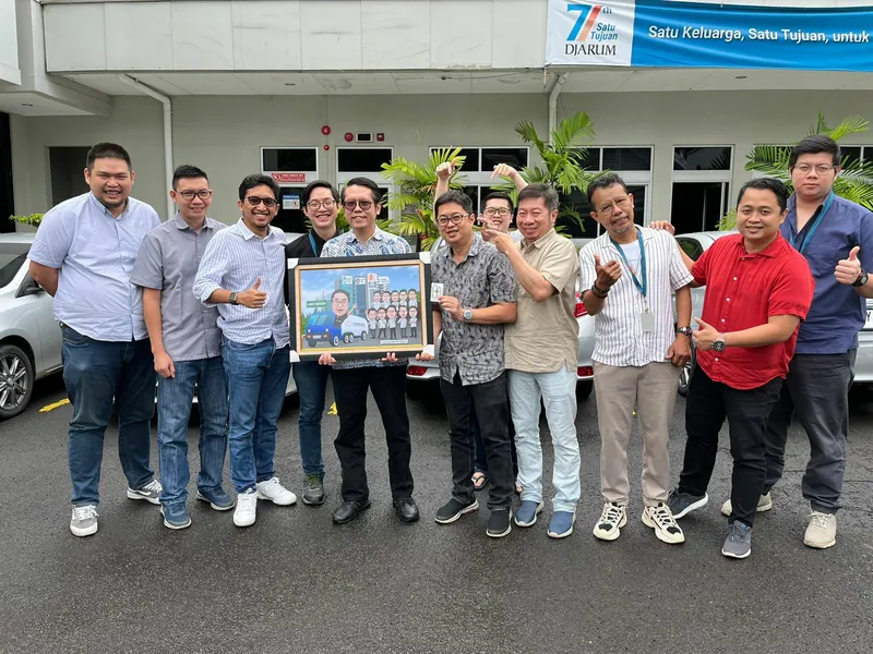 Rombongan rekan kerja berfoto di halaman kantor sambil memegang karikatur kelompok berbingkai.