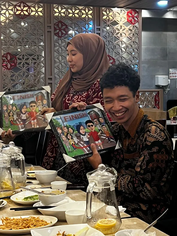 Perempuan berhijab dan rekan prianya memegang karikatur berbingkai di meja makan restoran.