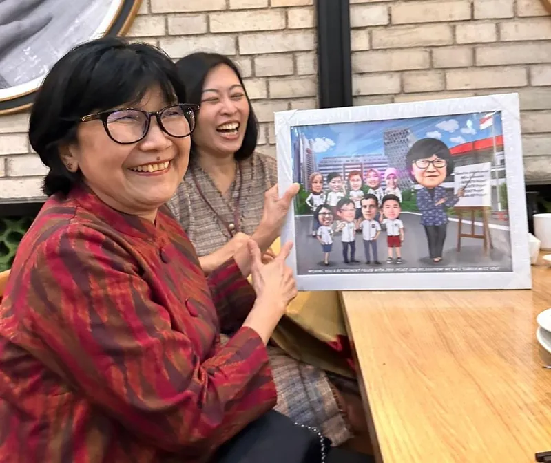 Dua perempuan tertawa sambil memegang karikatur kelompok berbingkai.