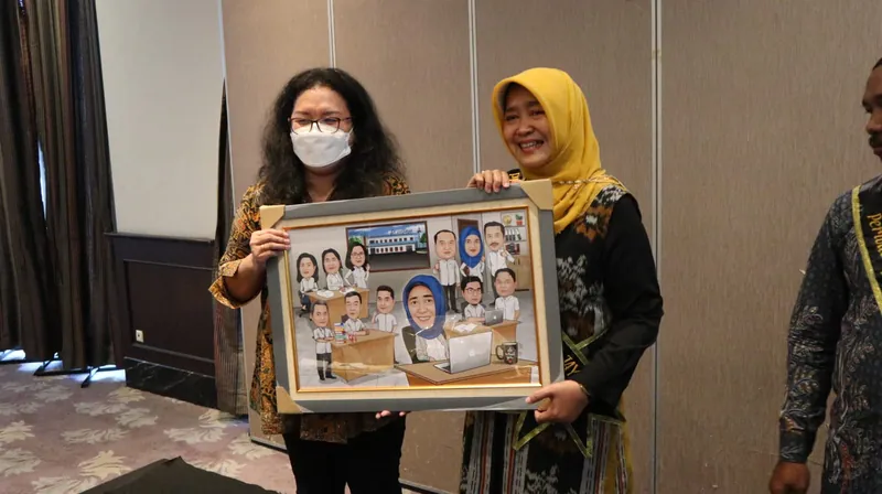 Dua perempuan memegang karikatur kelompok berbingkai di ruang acara.