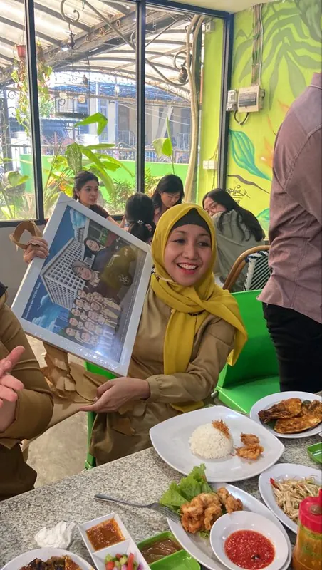 Perempuan berhijab kuning memegang karikatur berbingkai di meja makan.