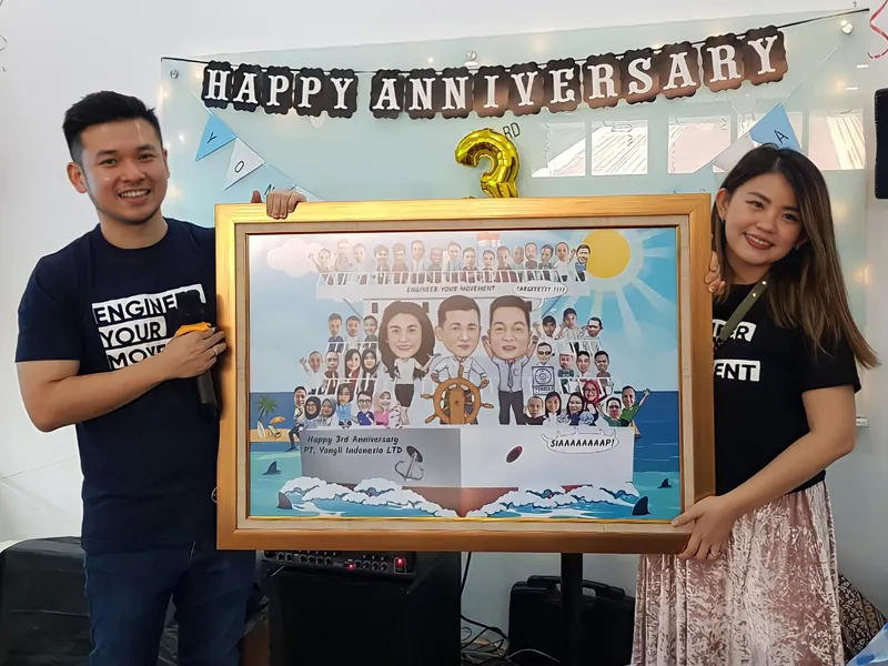 Dua orang memegang karikatur kelompok berbingkai besar di depan spanduk anniversary kantor.