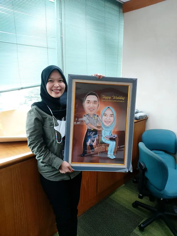 Perempuan berhijab memegang karikatur pasangan berbingkai di ruang kantor.