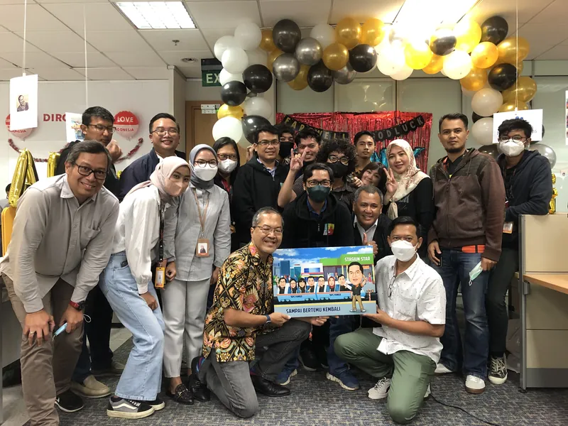 Karyawan berkumpul di ruang kerja berhias balon memegang karikatur kelompok berbingkai.