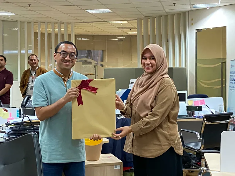 Penyerahan bingkisan dan karikatur berbingkai antara dua rekan kerja di ruang kantor.