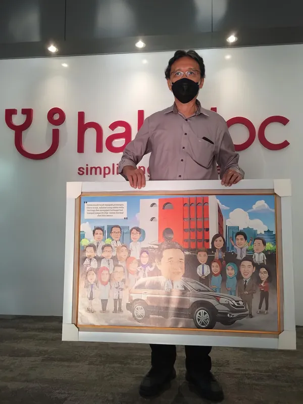 Seorang pria bermasker memegang karikatur kelompok berbingkai di kantor Halodoc.