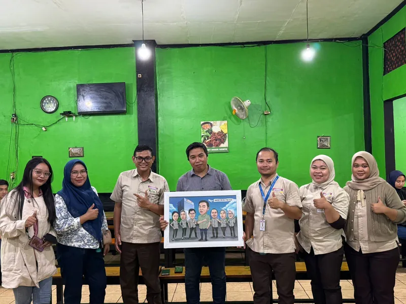 Pegawai berseragam cokelat memegang karikatur kelompok berbingkai di ruang berdinding hijau.