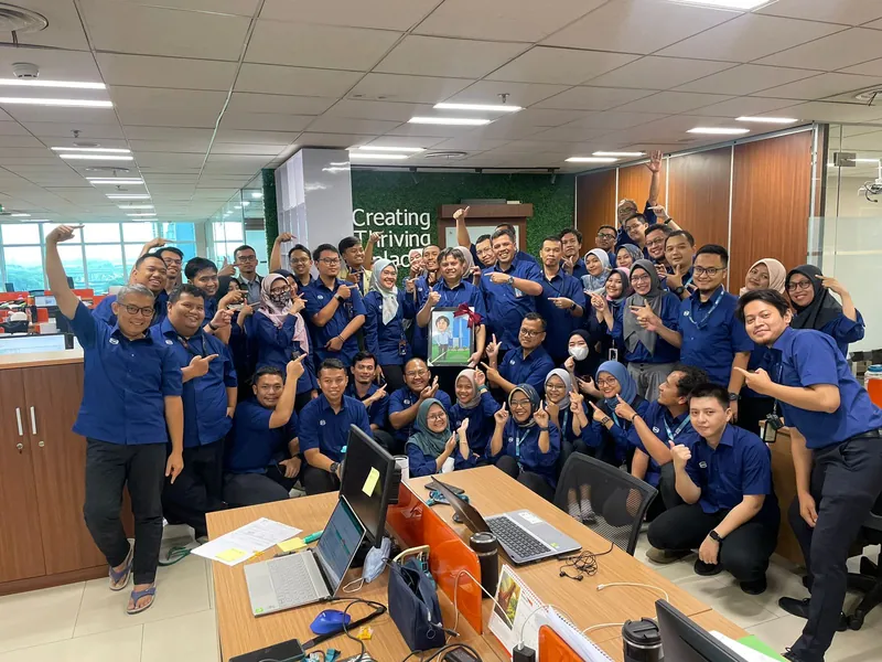 Karyawan berseragam biru merayakan penyerahan karikatur kelompok berbingkai di ruang kerja.
