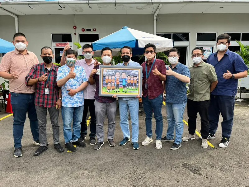 Rekan kerja berfoto di halaman kantor sambil memegang karikatur kelompok berbingkai.