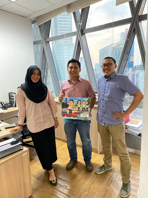 Tiga rekan kerja memegang karikatur kelompok berbingkai di ruang kantor berjendela tinggi.