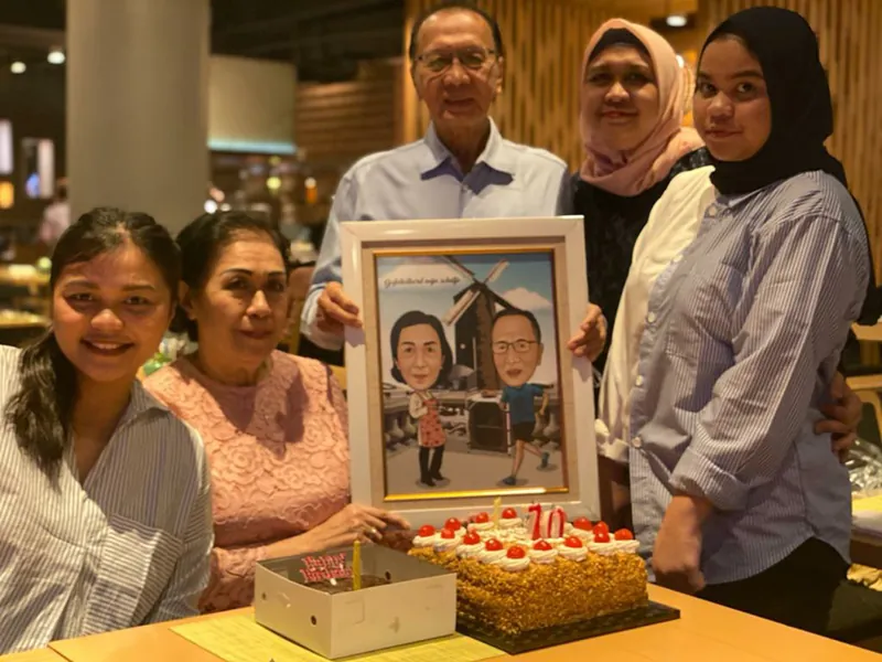 Rekan kerja berkumpul memegang karikatur berbingkai di samping kue ulang tahun.