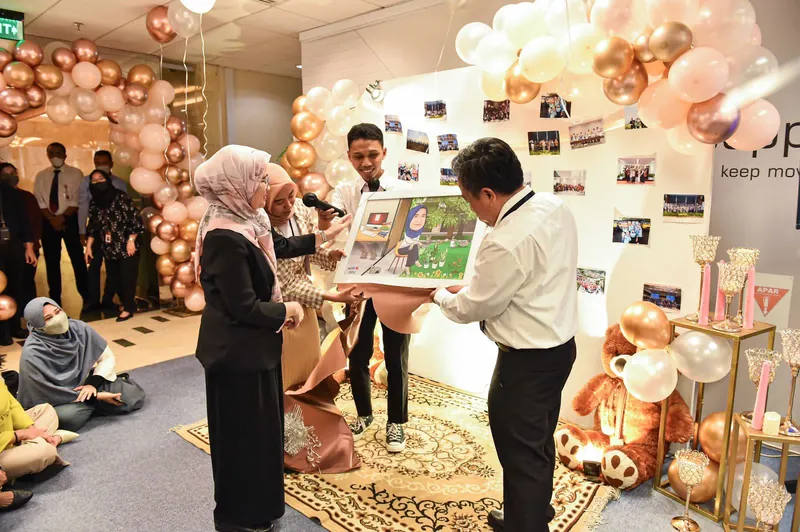Penyerahan karikatur berbingkai di ruang acara berhias balon dan dinding foto.