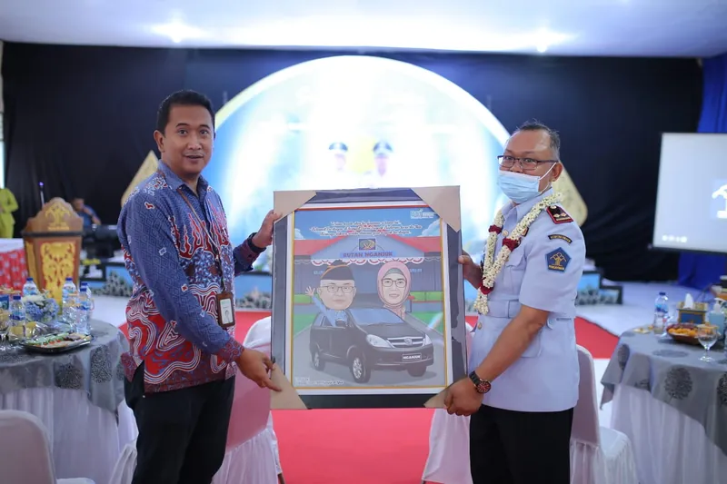 Penyerahan karikatur berbingkai bertema mobil kepada pejabat berseragam.