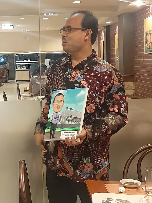 Seorang pria berbatik memegang karikatur berbingkai di ruang kerja.