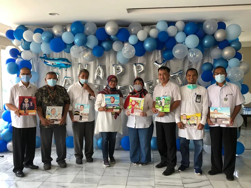 Karyawan berfoto di ruang berhias balon memegang karikatur berbingkai masing-masing.