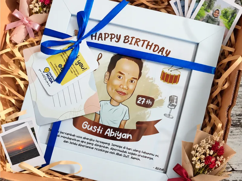 Karikatur berbingkai putih diikat pita biru di dalam kotak hampers berisi serutan kertas, buket bunga kering, kartu ucapan, dan cetakan foto polaroid